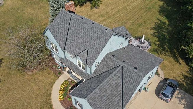 An aerial view of a light-blue two-story house with a grey shingled roof, a paved driveway, and a grassy backyard.