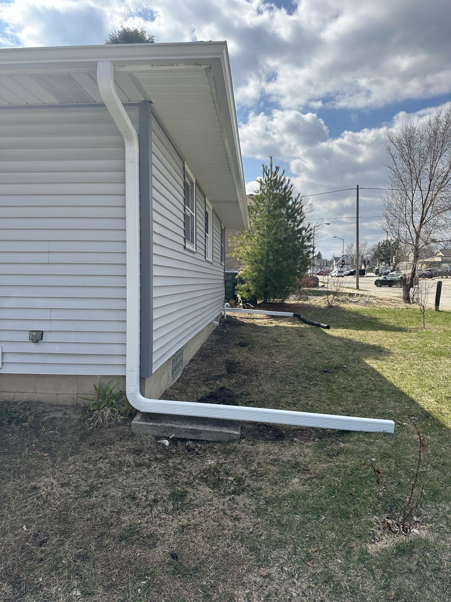 A white downspout extends from the corner of a light-sided house, resting on a concrete block in a grass yard.