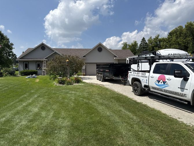 A white pickup truck with a large dump trailer parked on a gravel driveway in front of a gray, one-story house.