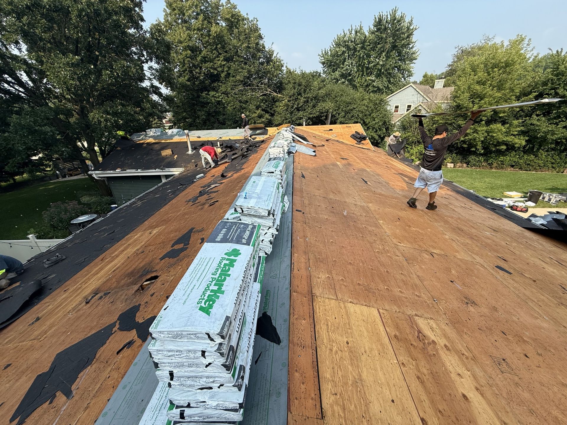 A worker repairs a roof with a stack of shingles along the ridge in a sunny outdoor setting.