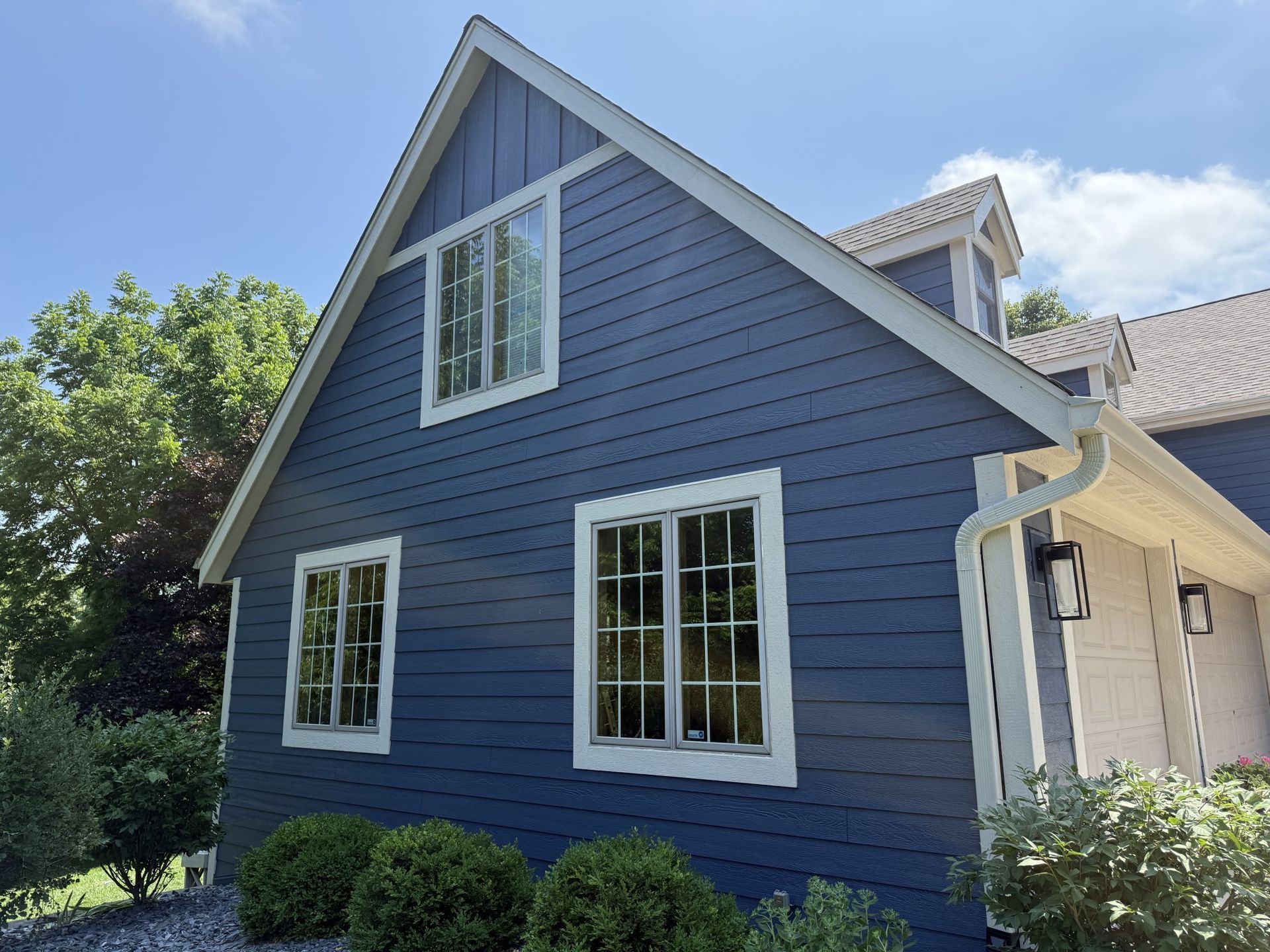 A blue, two-story house with white trim, large windows, and a steep roof, surrounded by green trees and bushes.