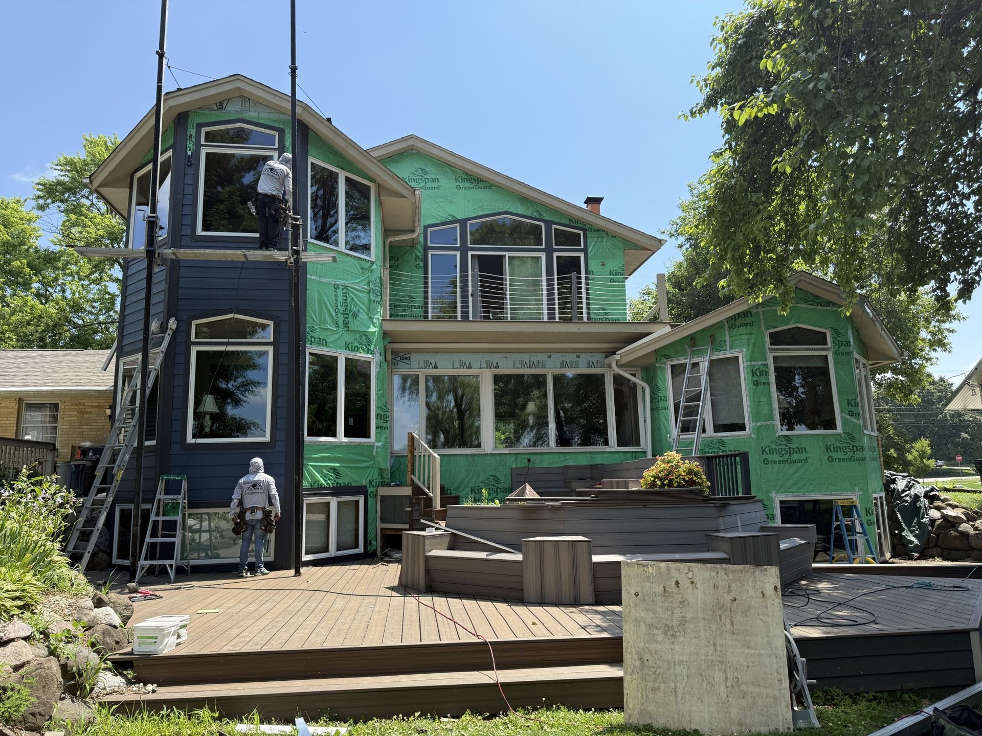 A two-story house under construction with green exterior sheathing, a wooden deck, and workers on scaffolding.