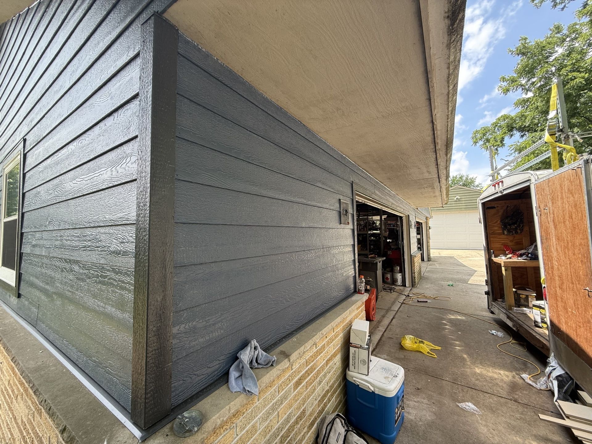 Newly painted dark gray siding on a house exterior next to a brick wall, with a trailer parked in the adjacent driveway.