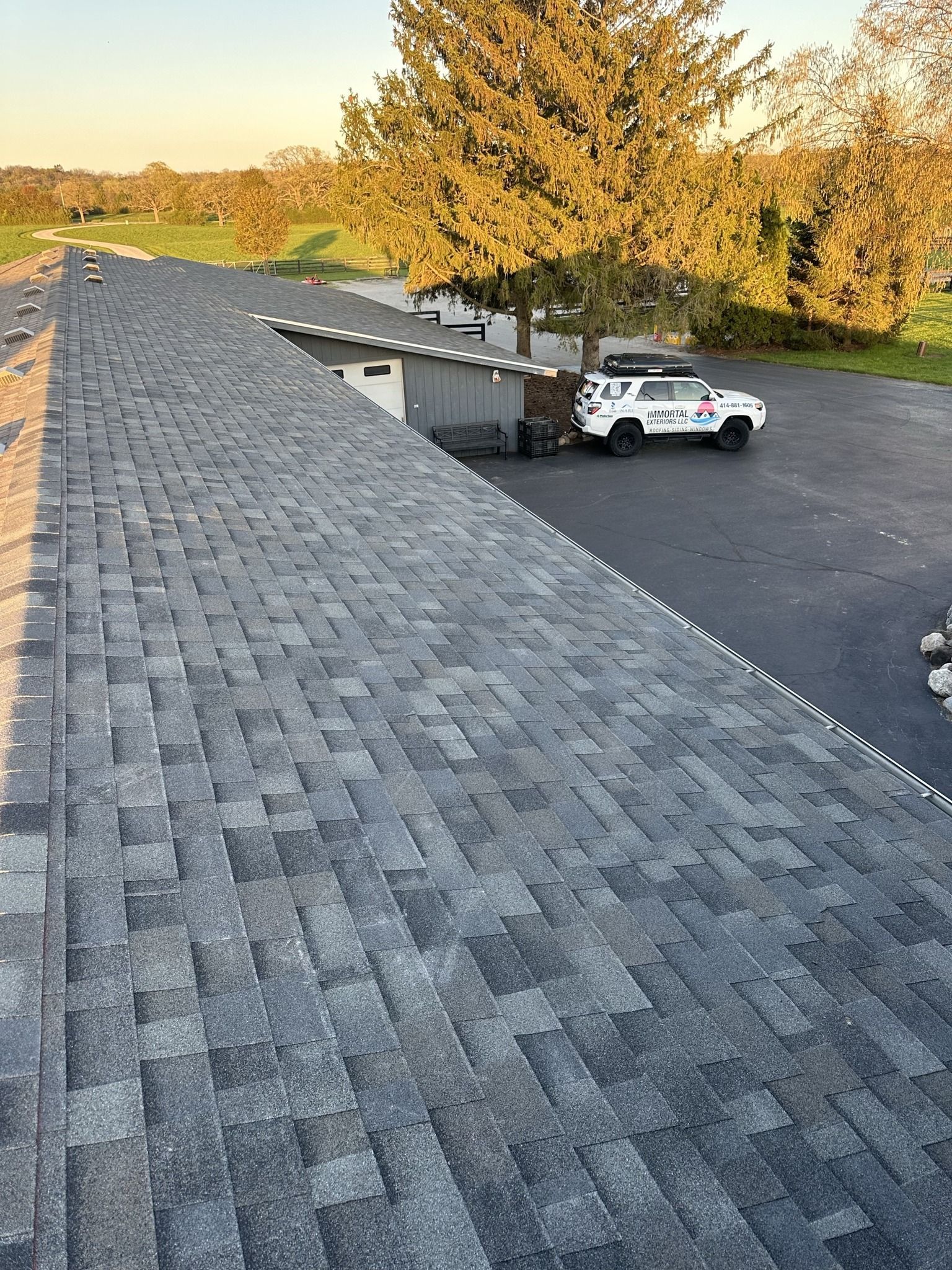 A high-angle view of a newly shingled gray roof, with a white vehicle parked in a paved driveway below.