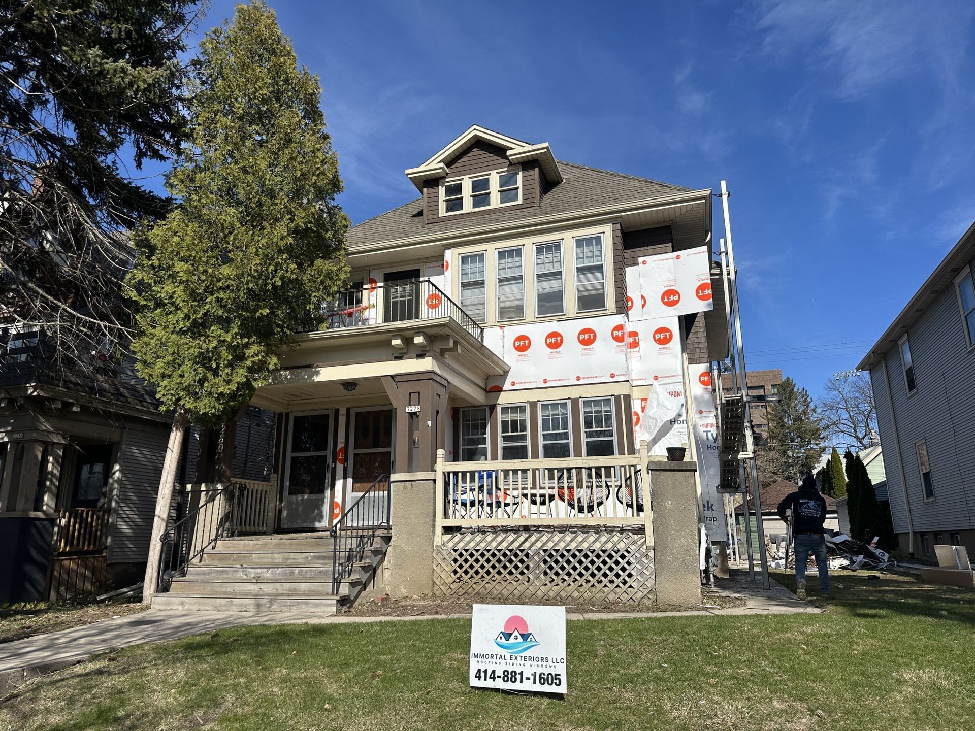 A two-story house under renovation with protective white siding wrap, a front porch, and a lawn sign in front.