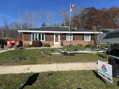 A single-story suburban home undergoing roof repairs, featuring a ladder, debris on the lawn, and an American flag.