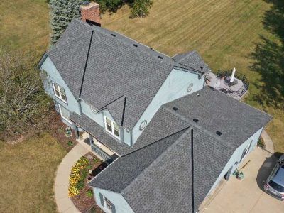 High-angle drone view of a blue house with a gray shingled roof, surrounded by a grassy lawn and a curved walkway.