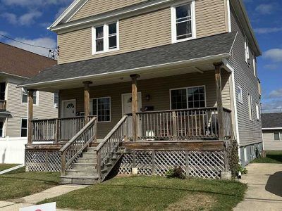 A two-story tan house with a large wooden front porch, lattice skirting, and a gabled roof under a blue, cloudy sky.