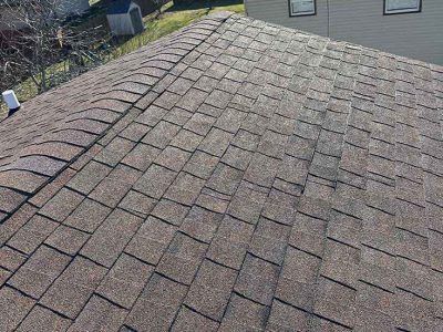 A high-angle view of a brown asphalt shingle roof, showing overlapping shingles and the ridgeline.