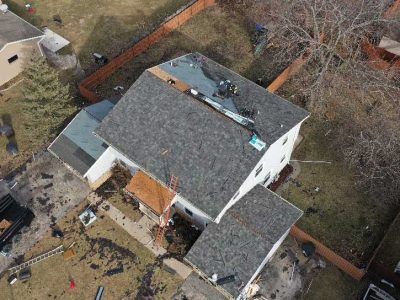 Aerial view of a house undergoing a roof replacement, with shingles scattered on the ground and scaffolding visible.
