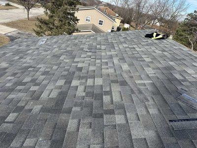 A high-angle view of a newly shingled gray asphalt roof, with a suburban home and trees visible in the background.