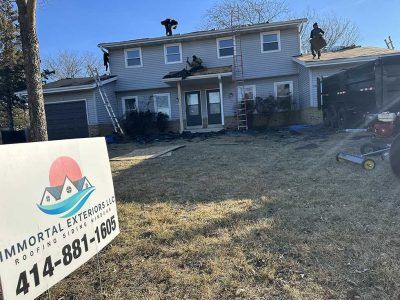 Contractors work on the roof of a two-story house with a lawn sign for Immortal Exteriors visible in the foreground.