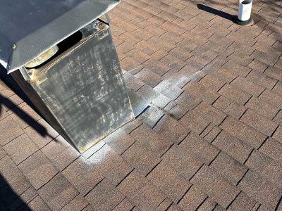 A close-up view of a metal roof vent flashing integrated into brown asphalt shingles on a sloped roof.