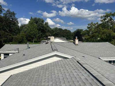 A view from above looking across gray shingled roofs toward tree-lined streets under a blue sky with scattered clouds.