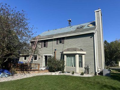 A construction worker on the roof of a gray two-story house with siding, performing repairs on a sunny day.