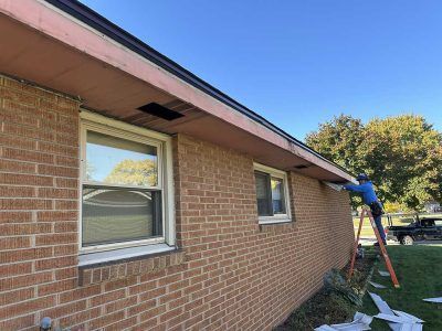 A person on a ladder repairs the soffit on the exterior of a tan brick house on a sunny day.