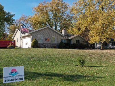 A tan house sits on a grassy hill with autumn trees, a red dumpster, and a sign with a phone number in the foreground.