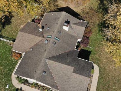 Aerial view of a residential house roof with shingles, a chimney, multiple skylights, and vent pipes, set on a lawn.
