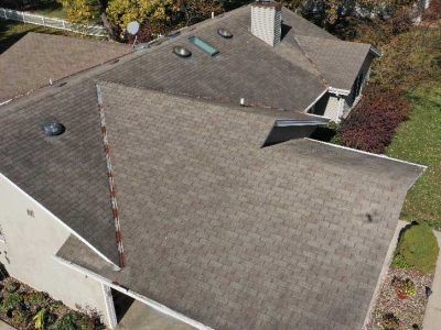 Aerial view of a residential shingled roof with a chimney, several skylights, and multiple roofline angles.