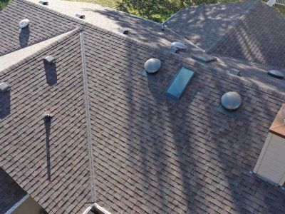 An aerial view of a dark grey shingled residential roof with multiple vents, a chimney, and a skylight.
