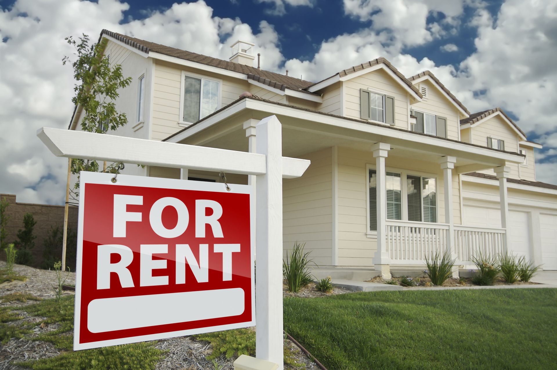 For rent sign in front of a beige house with a porch and cloudy sky