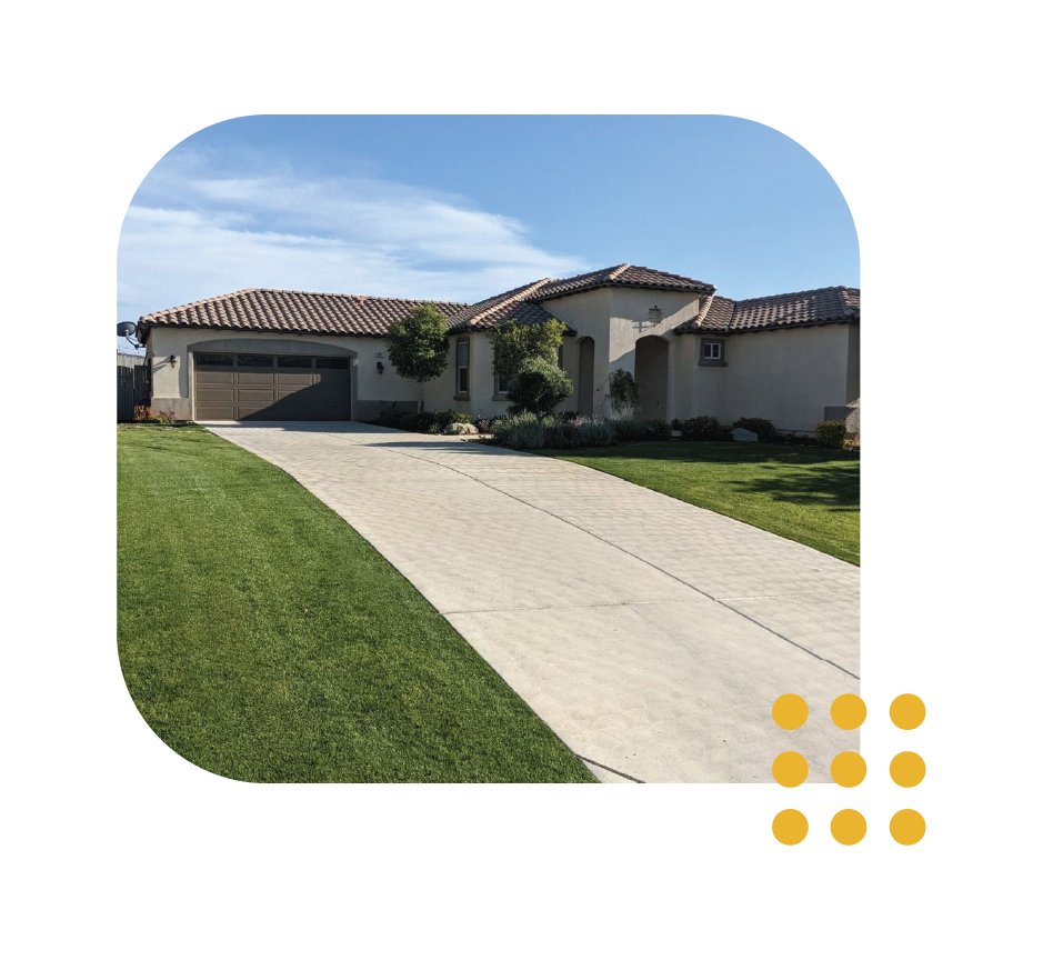 A single-story suburban house with a tan stucco exterior, a tile roof, a multi-car garage, and a long concrete driveway.