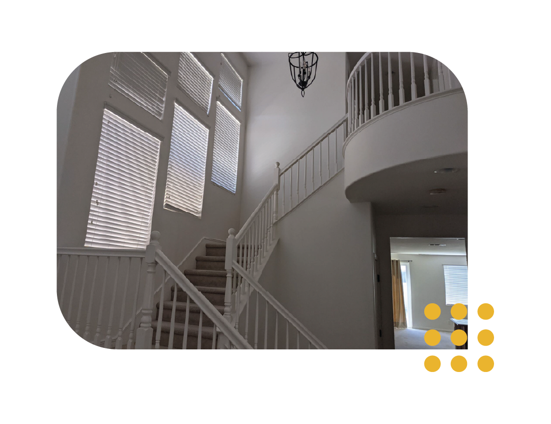 A white interior staircase with a curved landing, wood railings, and tall windows with blinds in a high-ceilinged foyer.