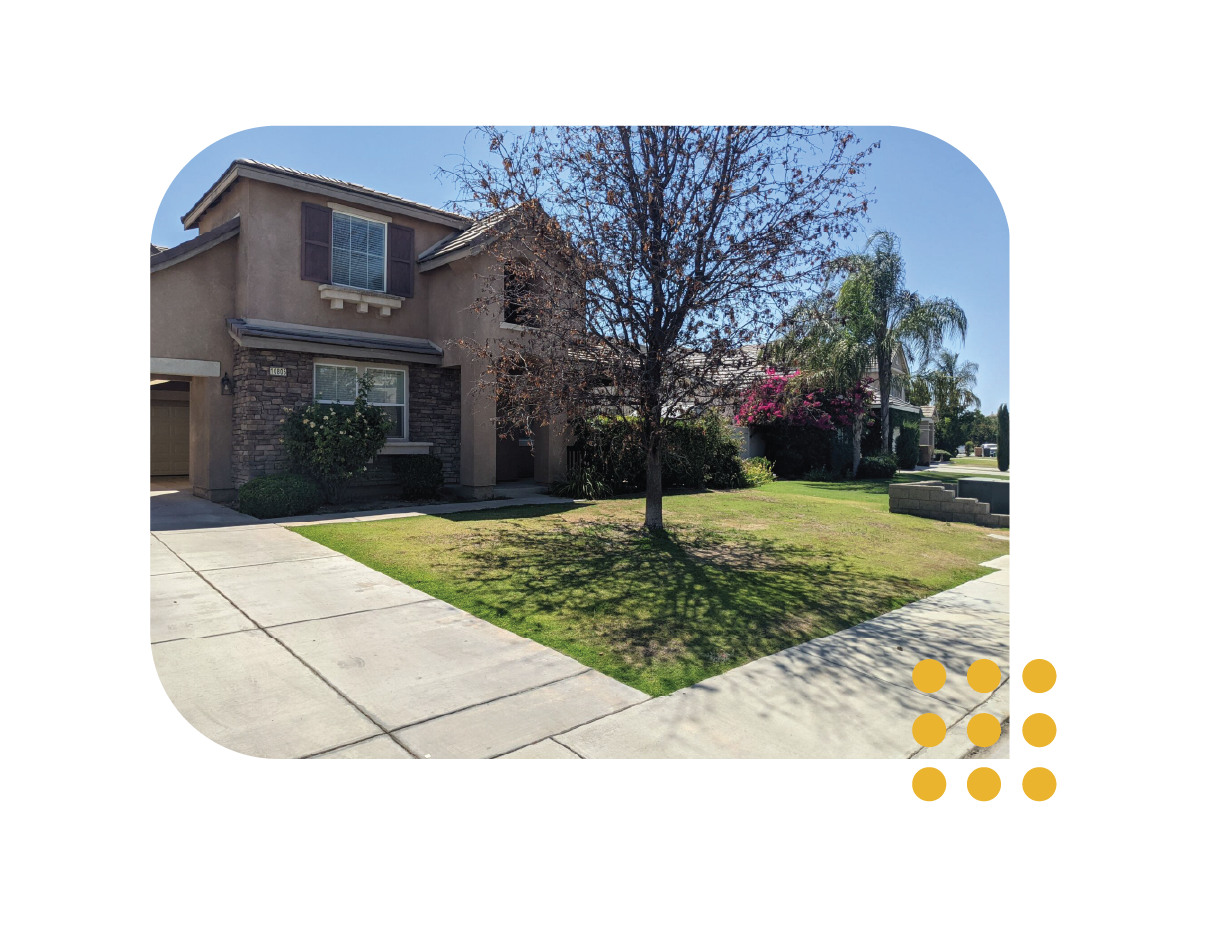 A two-story tan suburban house with a stone facade, a concrete driveway, and a front yard with a tree and bushes.