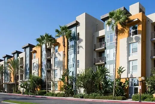 A multi-story apartment complex featuring tan and orange walls, glass-enclosed stairwells, balconies, and palm trees.