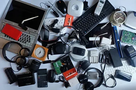 A top-down view of various electronic waste, including a laptop, headphones, keyboards, cameras, and cords on a white floor.