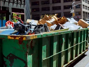 A green dumpster filled with cardboard boxes, white plastic bags, and discarded metal items on a city street.