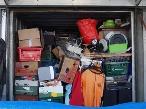 A storage unit filled with stacked cardboard boxes, plastic crates, a red bag, and various household items.