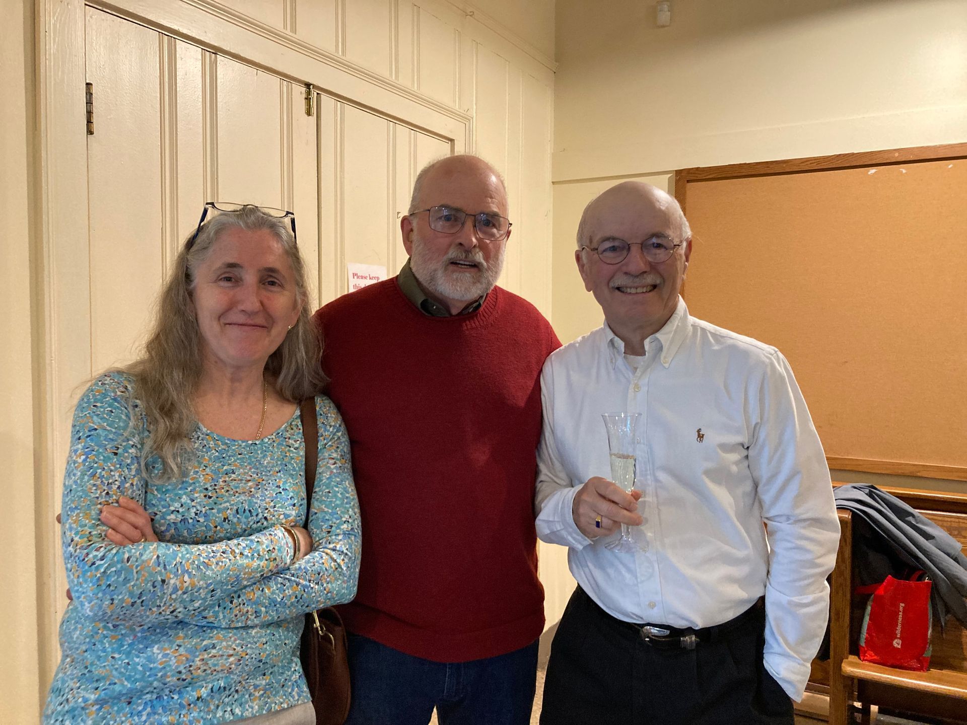 Three people smiling, indoors. Woman in blue top, man in red sweater, man in white shirt holding glass.