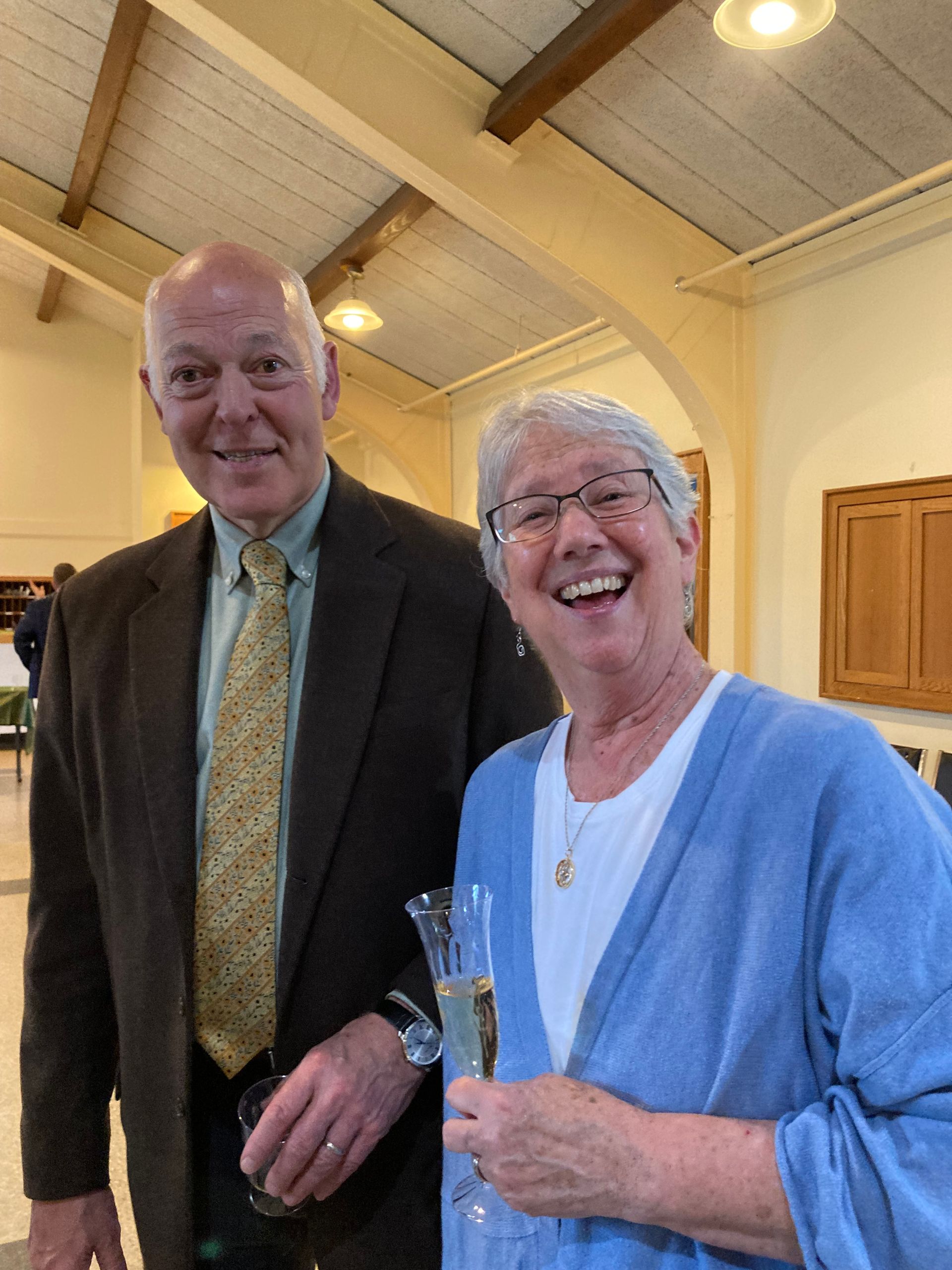 Man and woman smiling, indoors. Man in suit, woman holds champagne flute, blue cardigan.