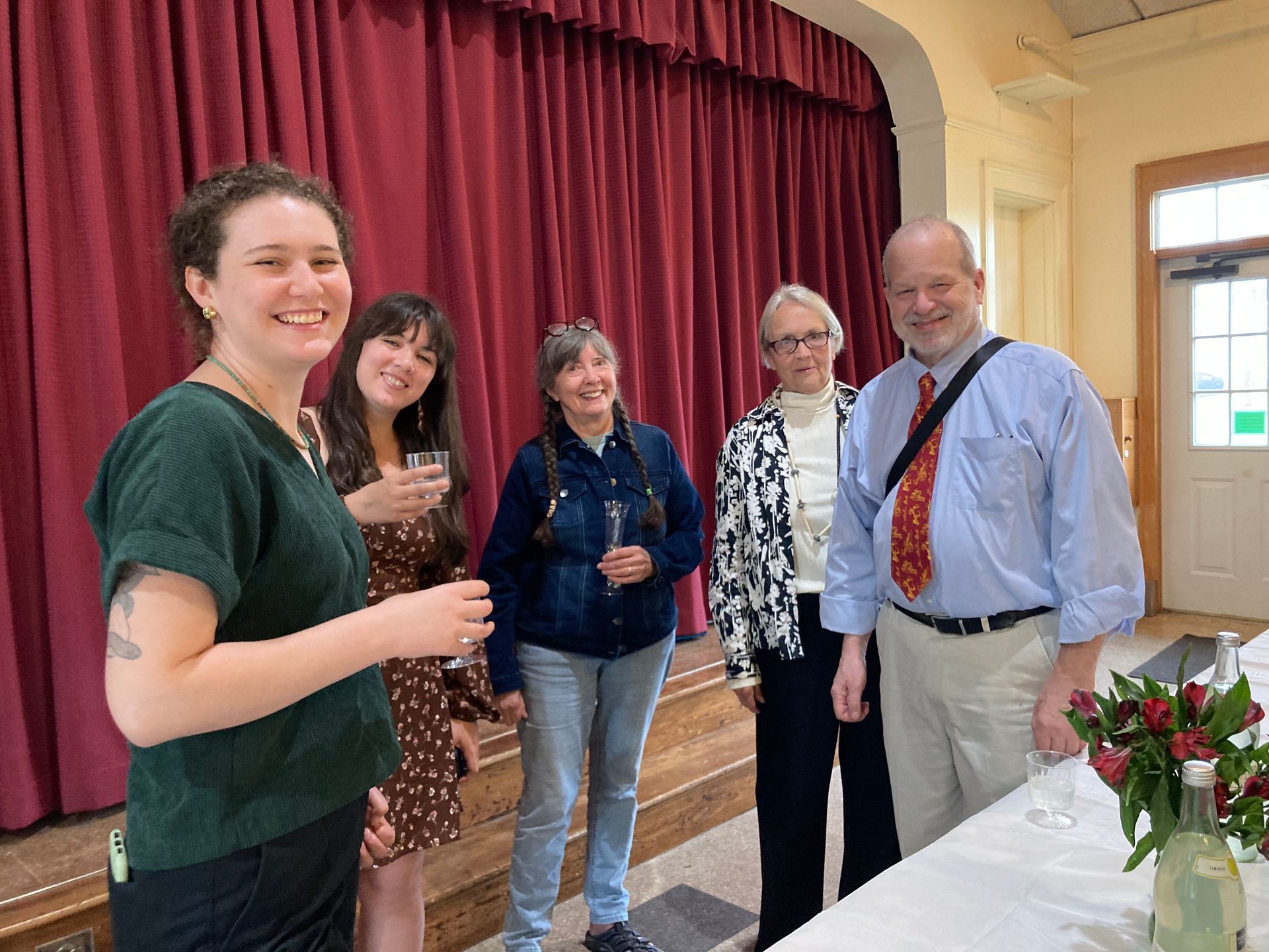 Group of six people smiling, standing in front of a red curtain, likely at an event.