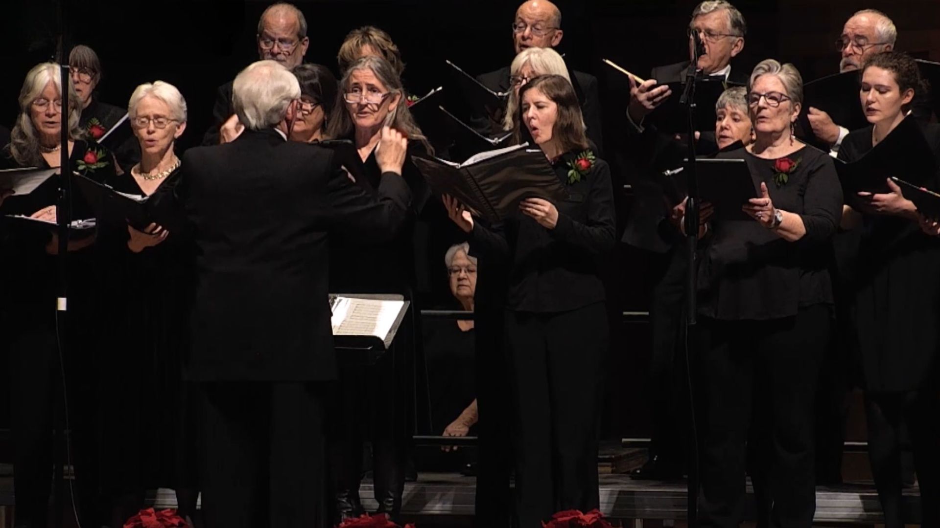 Choir singing, conductor leading. Stage lit with dark background, performers in black clothing, holding sheet music.