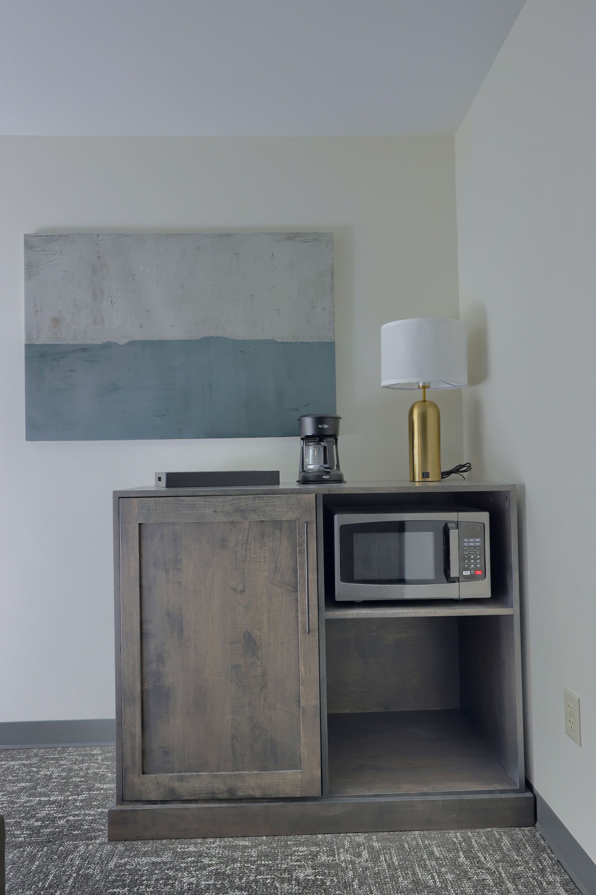 A room with a microwave , coffee maker , lamp and a painting on the wall.