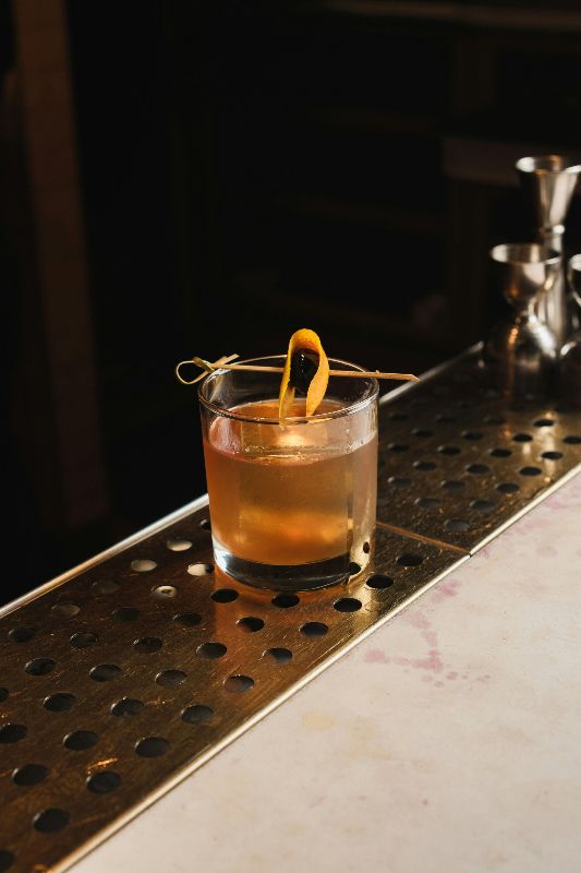 A glass of whiskey with an orange peel on a bar.