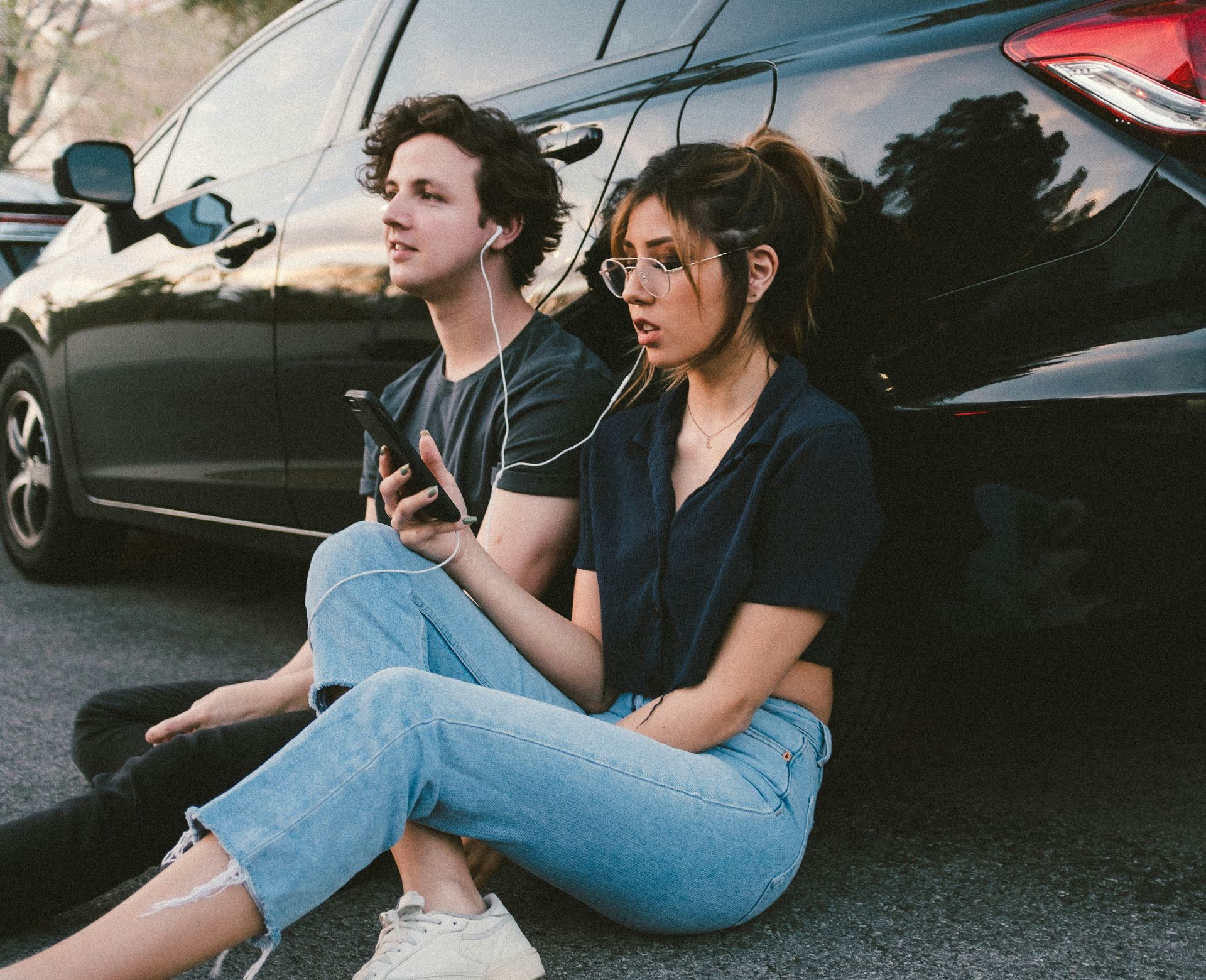 A man and woman sit on the road with their backs against a black car, listening to audio on a phone
