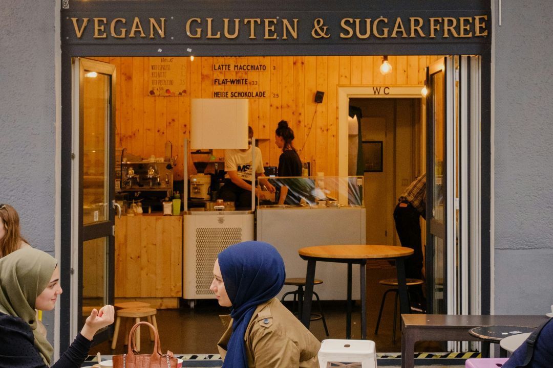 Two women in headscarves sit outside a coffee shop showing a sign for 'vegan gluten & sugar free'
