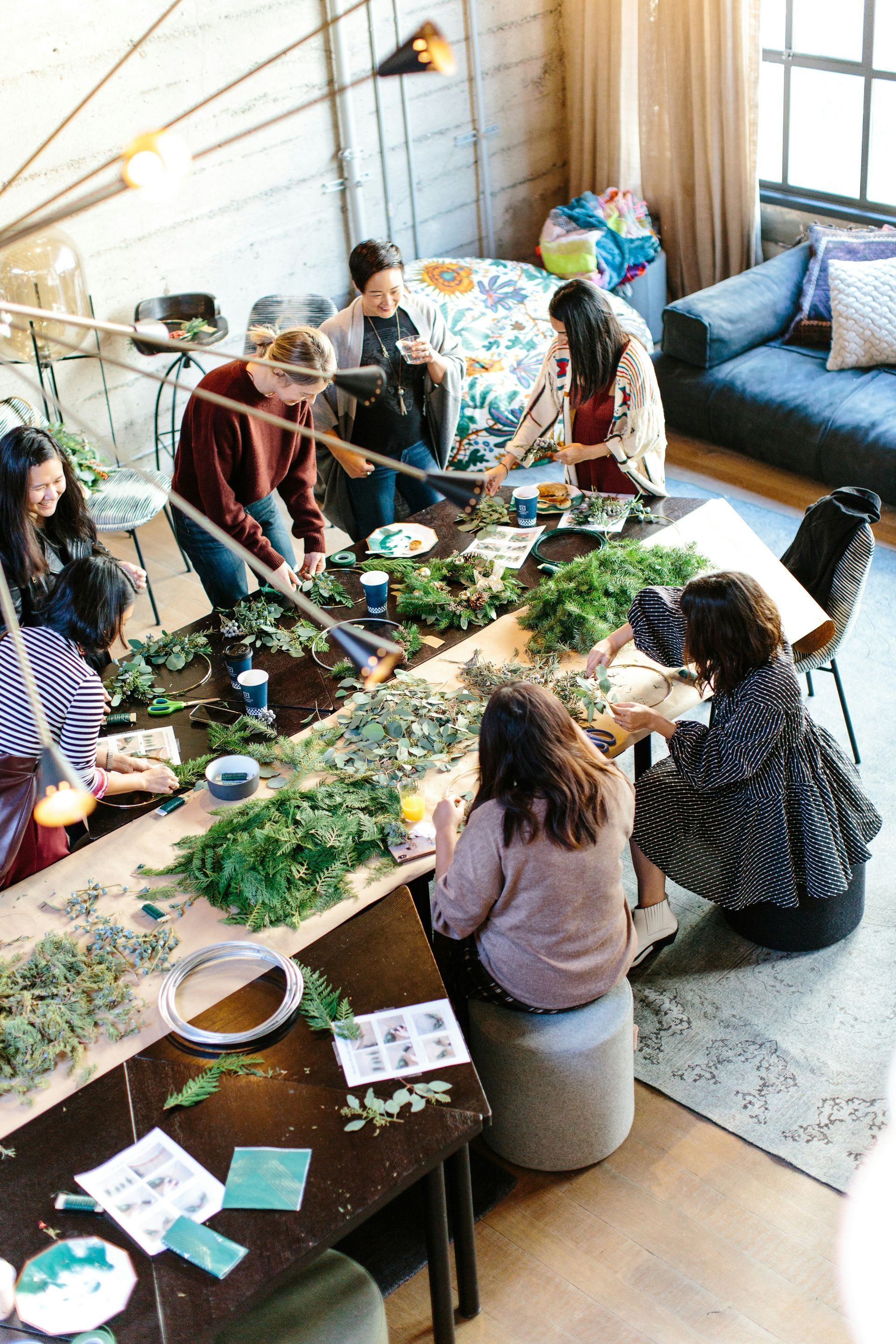 A group of women are gathered around a large table, constructing wreaths with plants and flowers.