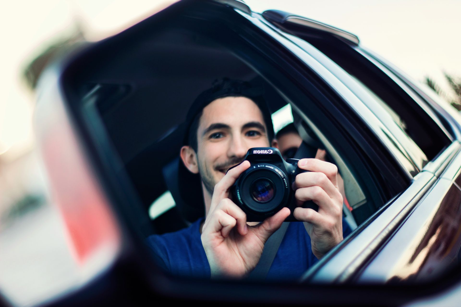 A man is taking a photo of himself in a car wing mirror using a Canon camera.