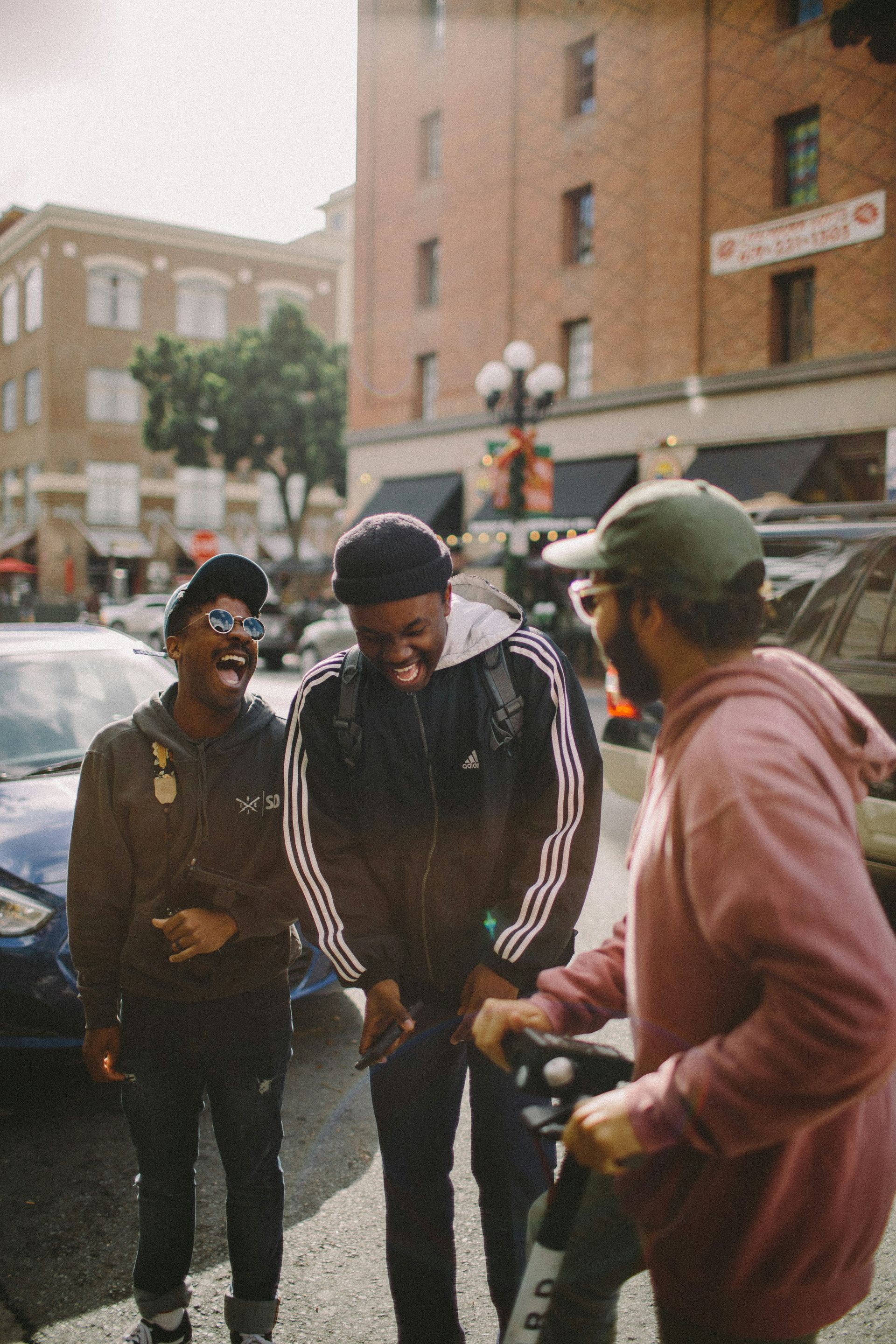 Three men dressed in casual clothes, caps, and sunglasses, are stood on the street laughing.