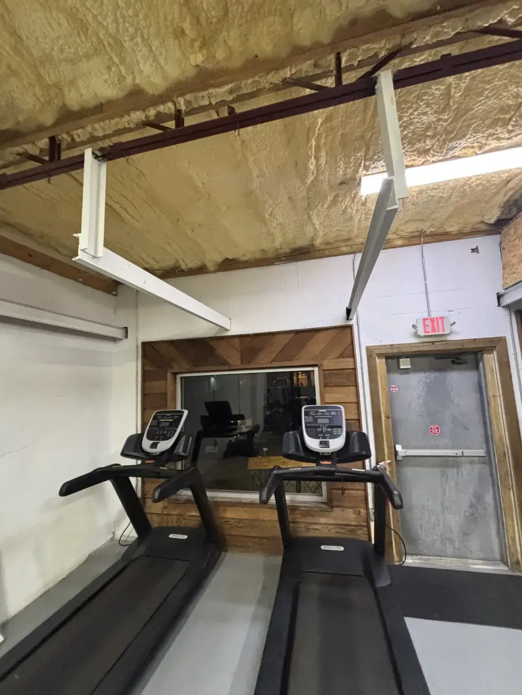 Two treadmills in a gym. Ceiling beams and insulation, window with wood paneling, and an exit door.