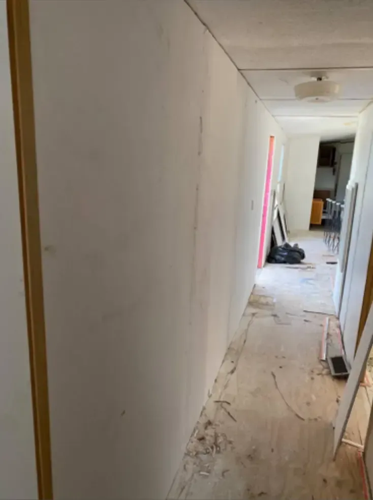 A hallway with white drywall, unfinished floors, and a view towards a room.