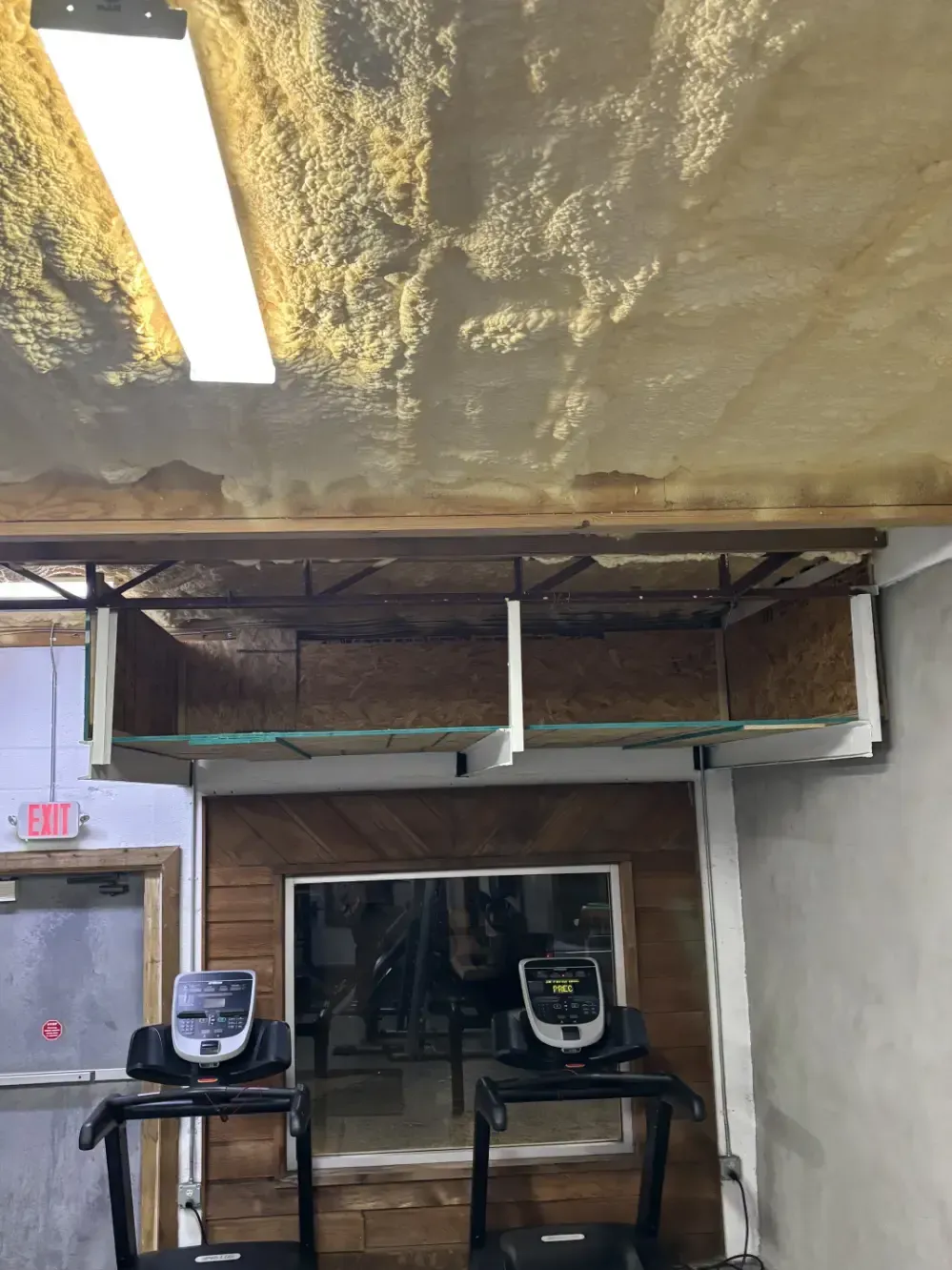 Gym with treadmills, mirror, and ceiling with exposed insulation and wood framing.