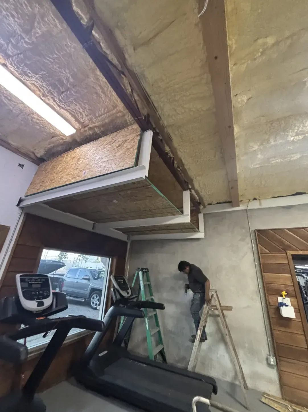 Man on ladder works on ceiling with wood beams. Treadmill, room with window, and wood paneling.
