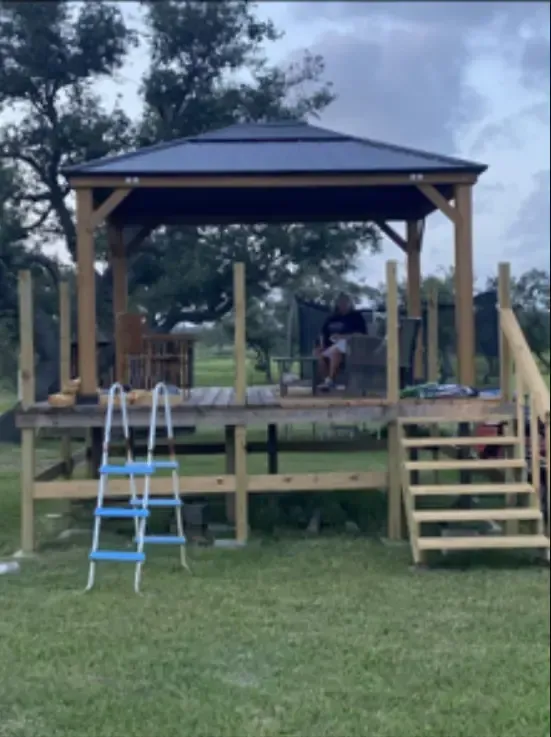 Gazebo on a wooden deck, person sitting inside. Blue ladder nearby. Green grass.