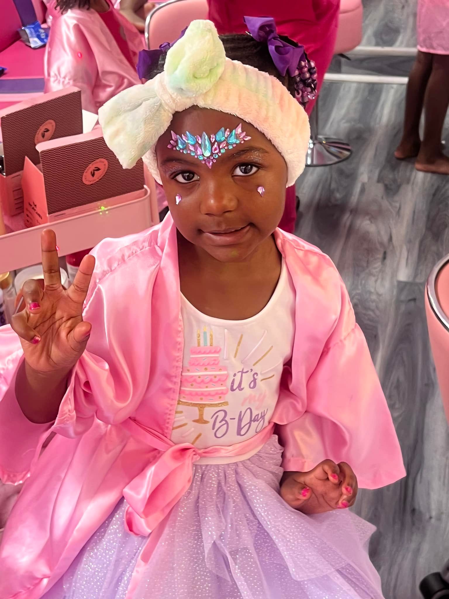 Girl celebrating birthday, wearing pink robe and tutu, peace sign, face paint.