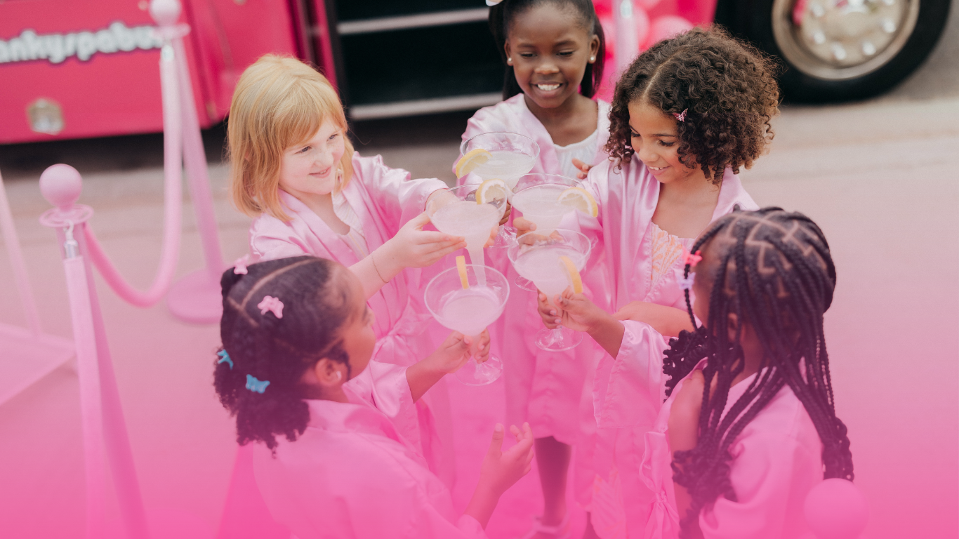 A group of young girls are toasting with drinks in front of a pink bus.